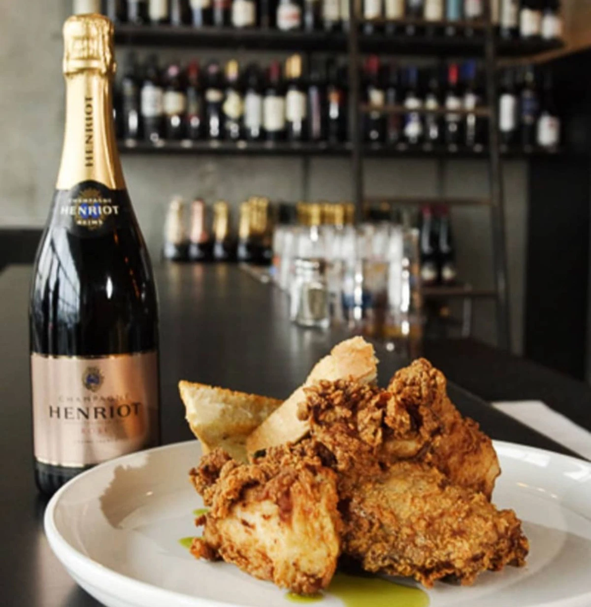 Henriot champagne bottle beside fried chicken on a white plate, with wine shelves behind
