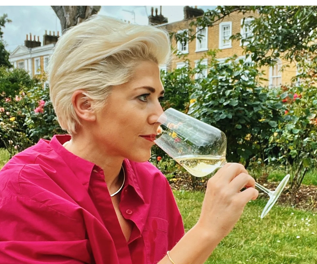 Woman in pink shirt smelling white wine in a garden with London houses behind her