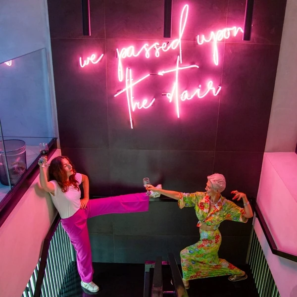 Two women holding wine glasses beneath pink neon sign reading we passed you the fair