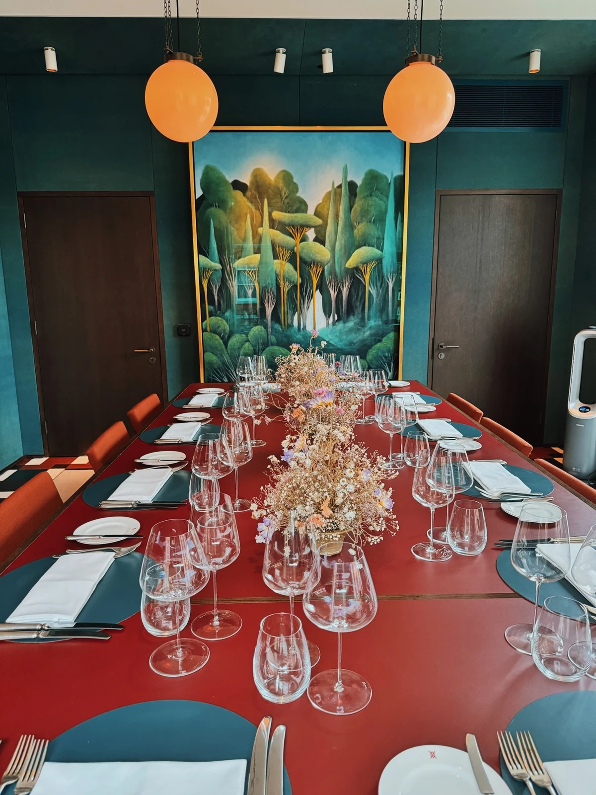 Long red dining table set with wine glasses, floral centrepiece and forest painting