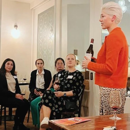 Person in orange speaking to seated women while holding a wine bottle and glass