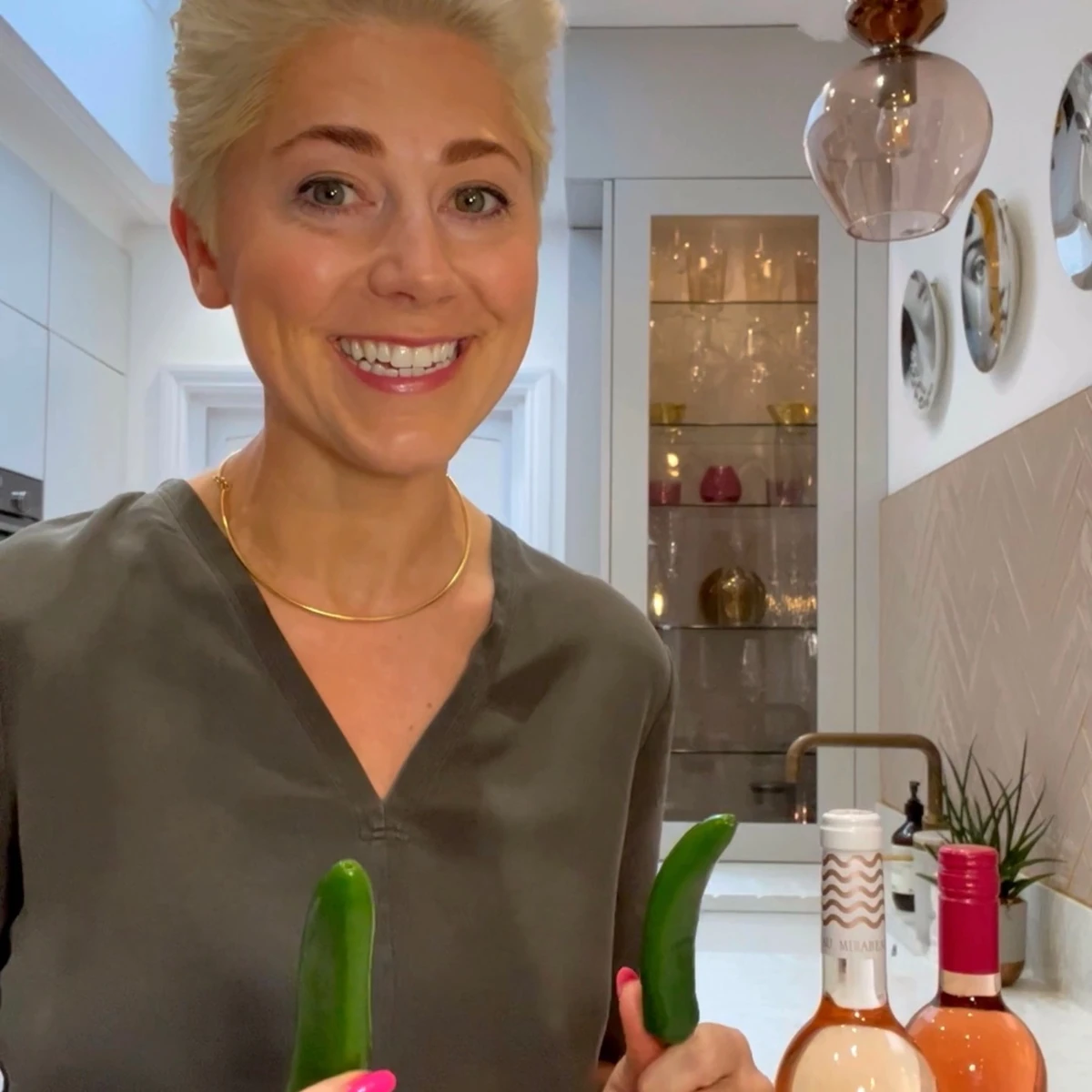 Smiling woman holding two cucumbers beside bottles of wine in a kitchen
