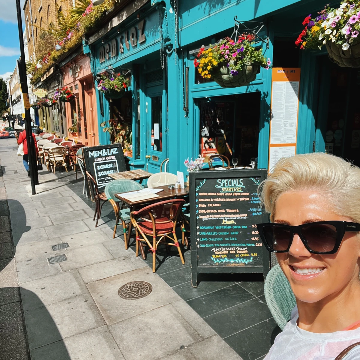 Woman in sunglasses beside turquoise restaurant with flower baskets and outdoor tables