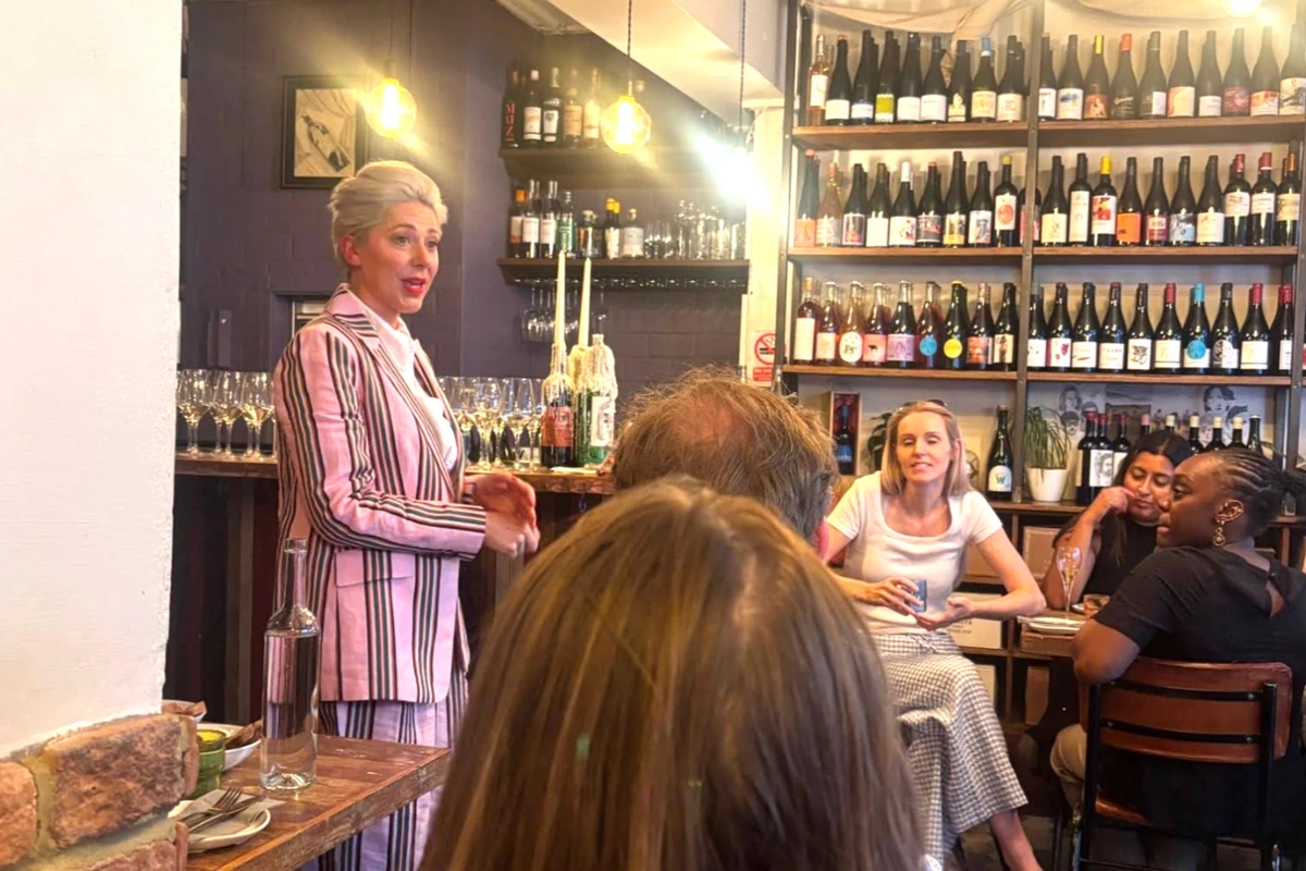 Wine tasting event with presenter, seated guests and shelves of wine bottles