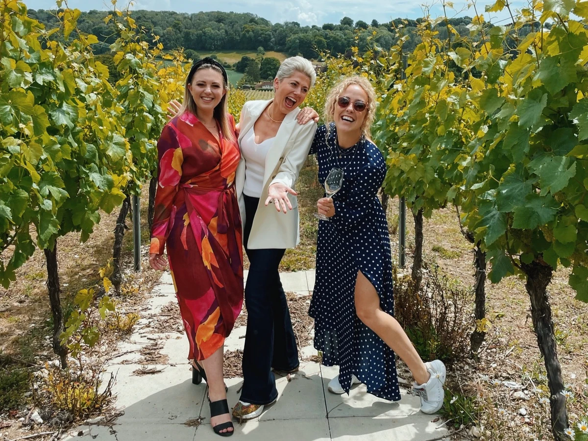 Three women posing among grapevines, one holding a wine glass