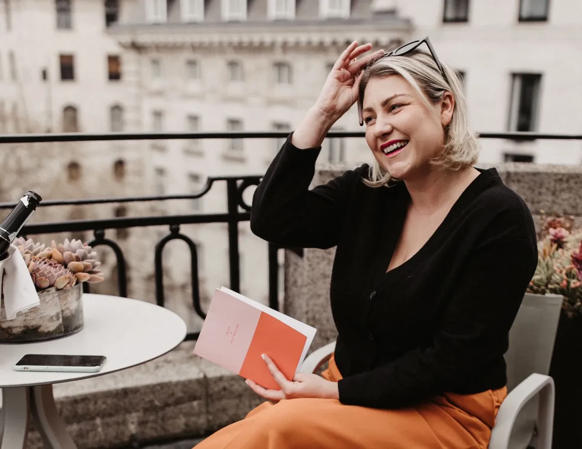 Smiling woman holding a pink and orange booklet on a balcony beside flowers