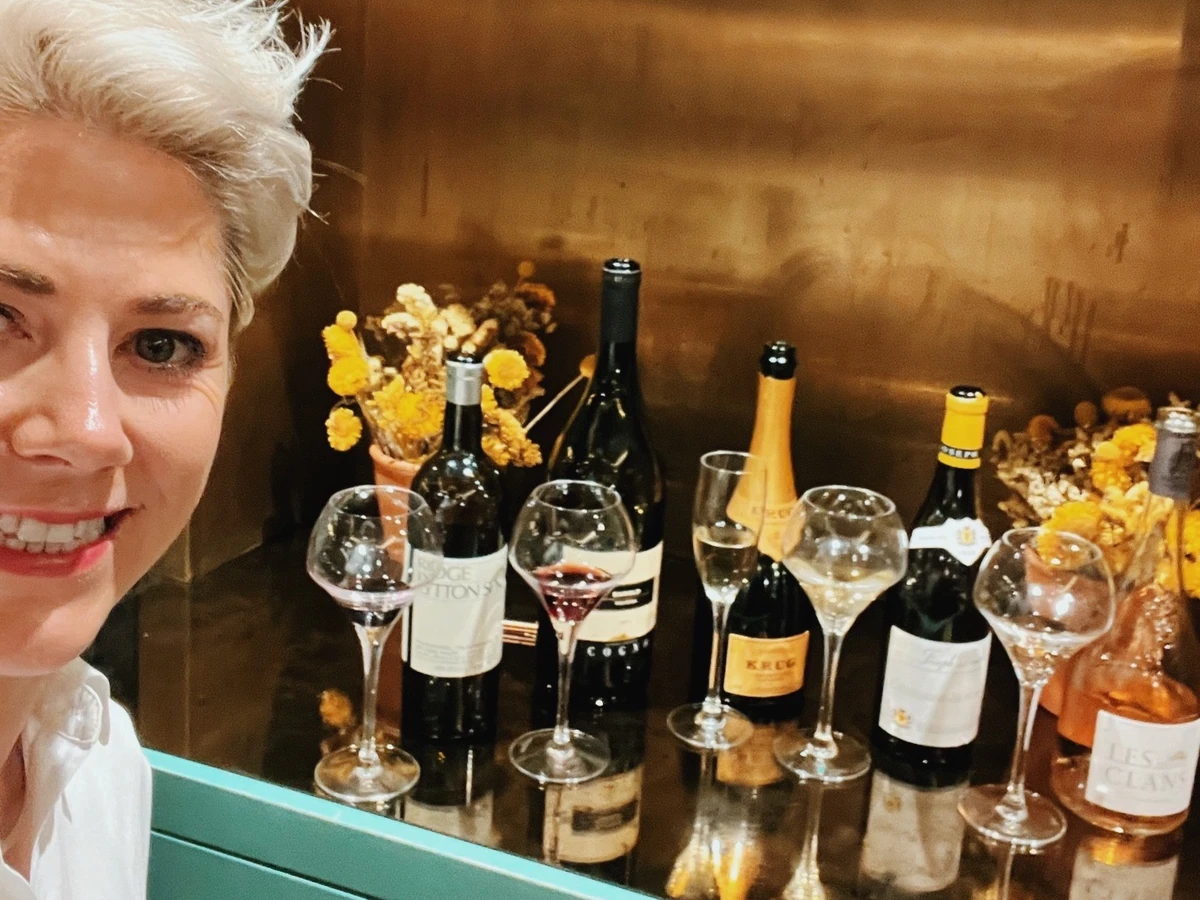 Person beside wine bottles and glasses, with dried flowers on a shelf