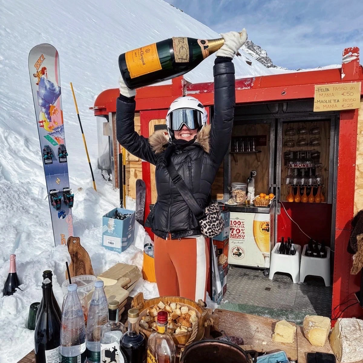 Skier holding a large champagne bottle outside a snowy mountain hut