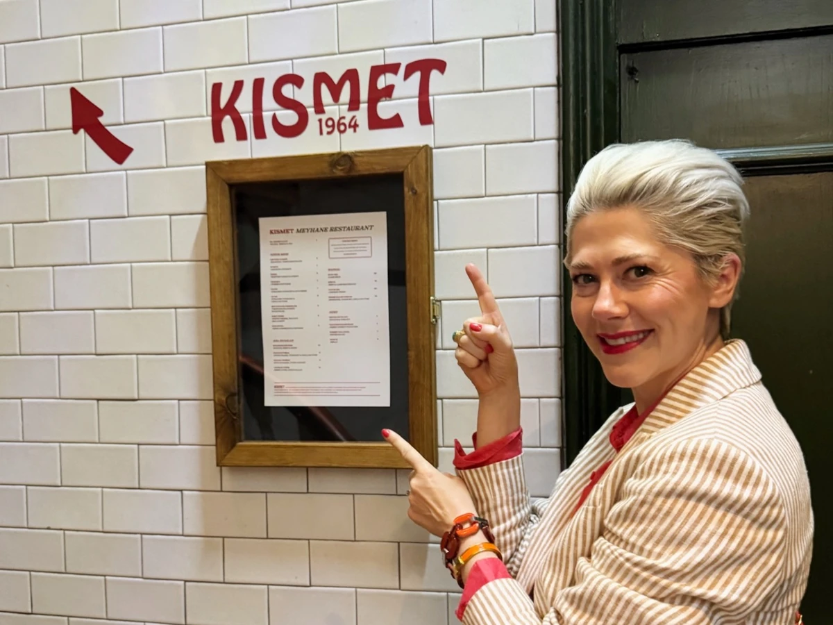 Woman pointing to KISMET 1964 restaurant sign and framed menu on tiled wall