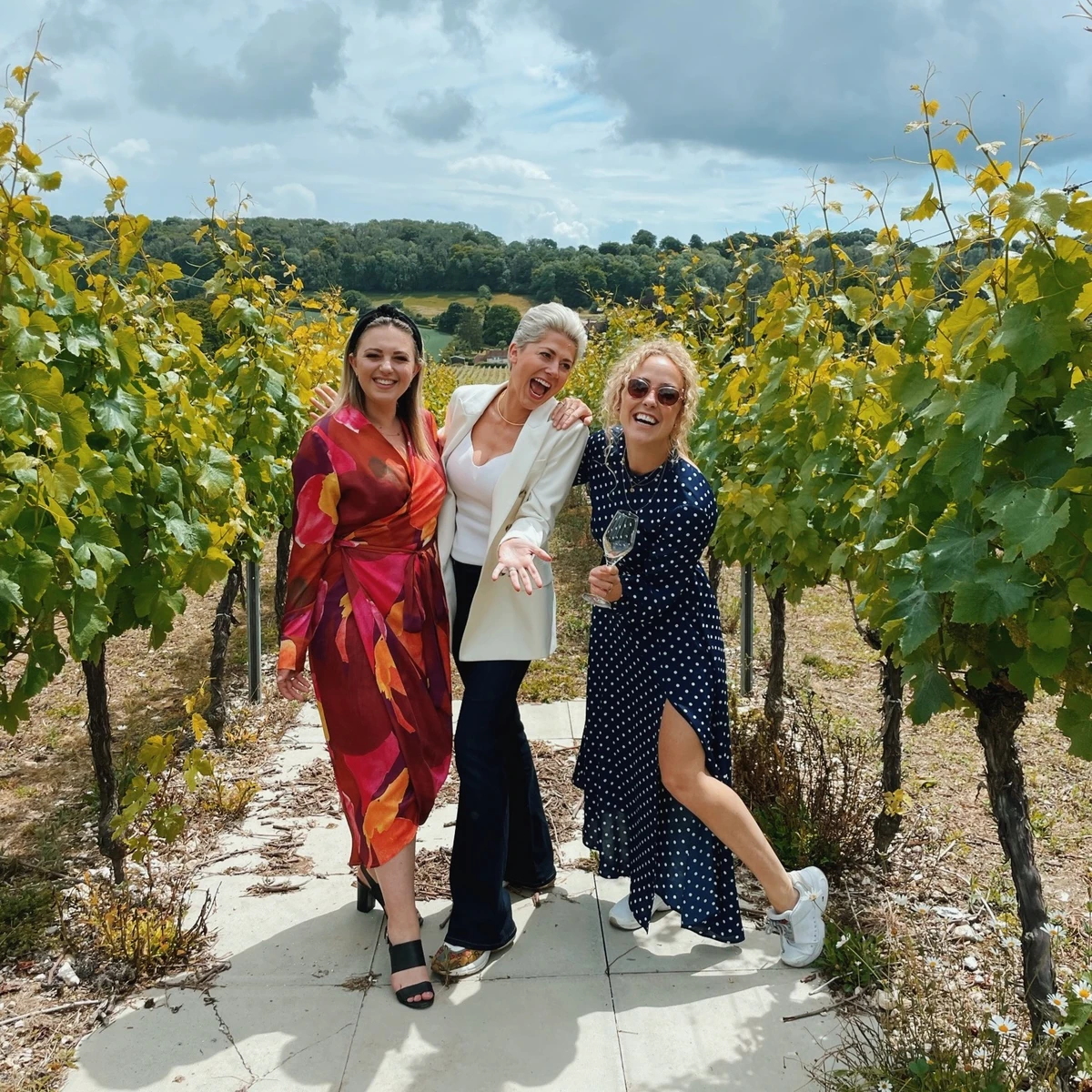 Three women posing among vineyard rows under a cloudy sky