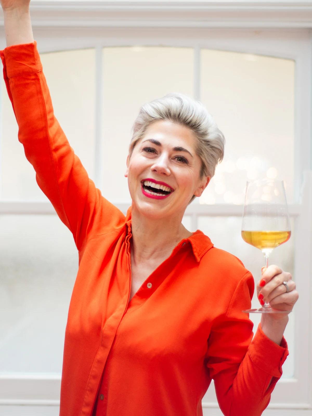Smiling woman in bright orange shirt raising her arm while holding a glass of white wine