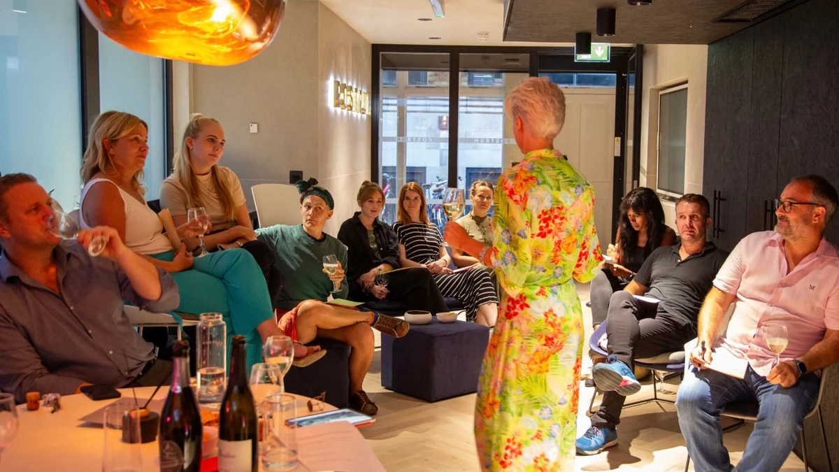 People seated indoors listening to a speaker in a colourful floral outfit, with wine bottles visible