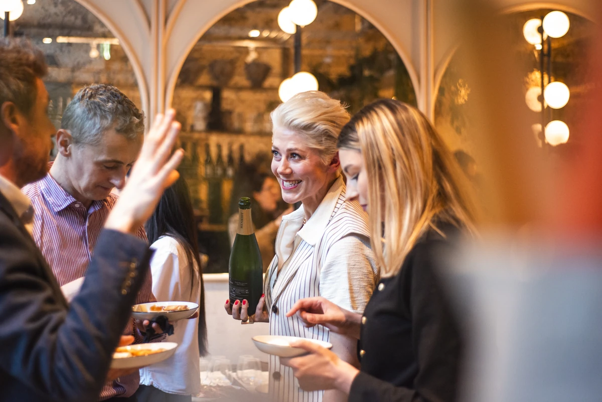 People mingling at an indoor event, with a woman holding a wine bottle and glasses