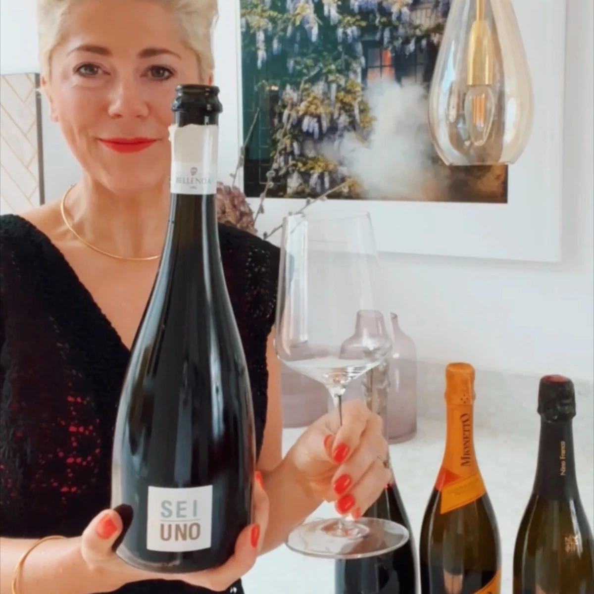 Woman holding a bottle labelled SEI UNO beside two sparkling wine bottles and glasses