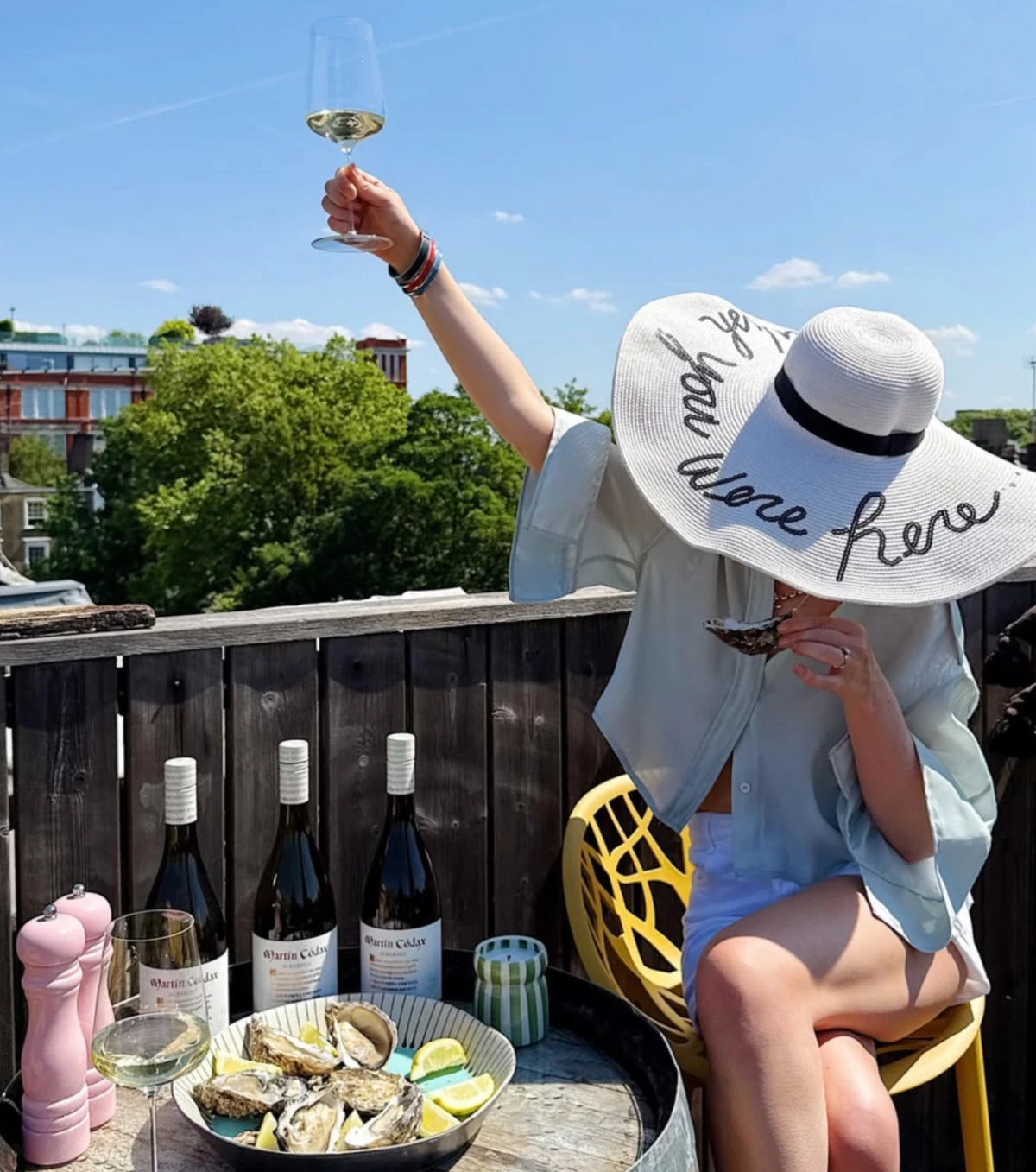 Person raising a wine glass beside oysters and wine bottles on a sunny rooftop