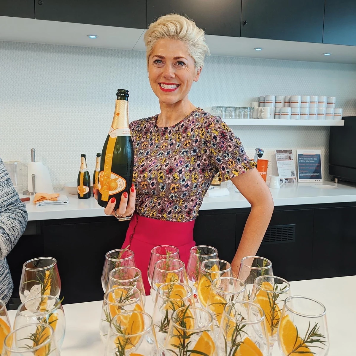 Smiling woman holding a champagne bottle behind glasses garnished with orange slices and rosemary