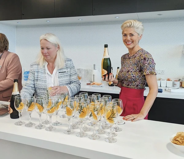 Two women serve sparkling wine with orange garnishes at a kitchen counter