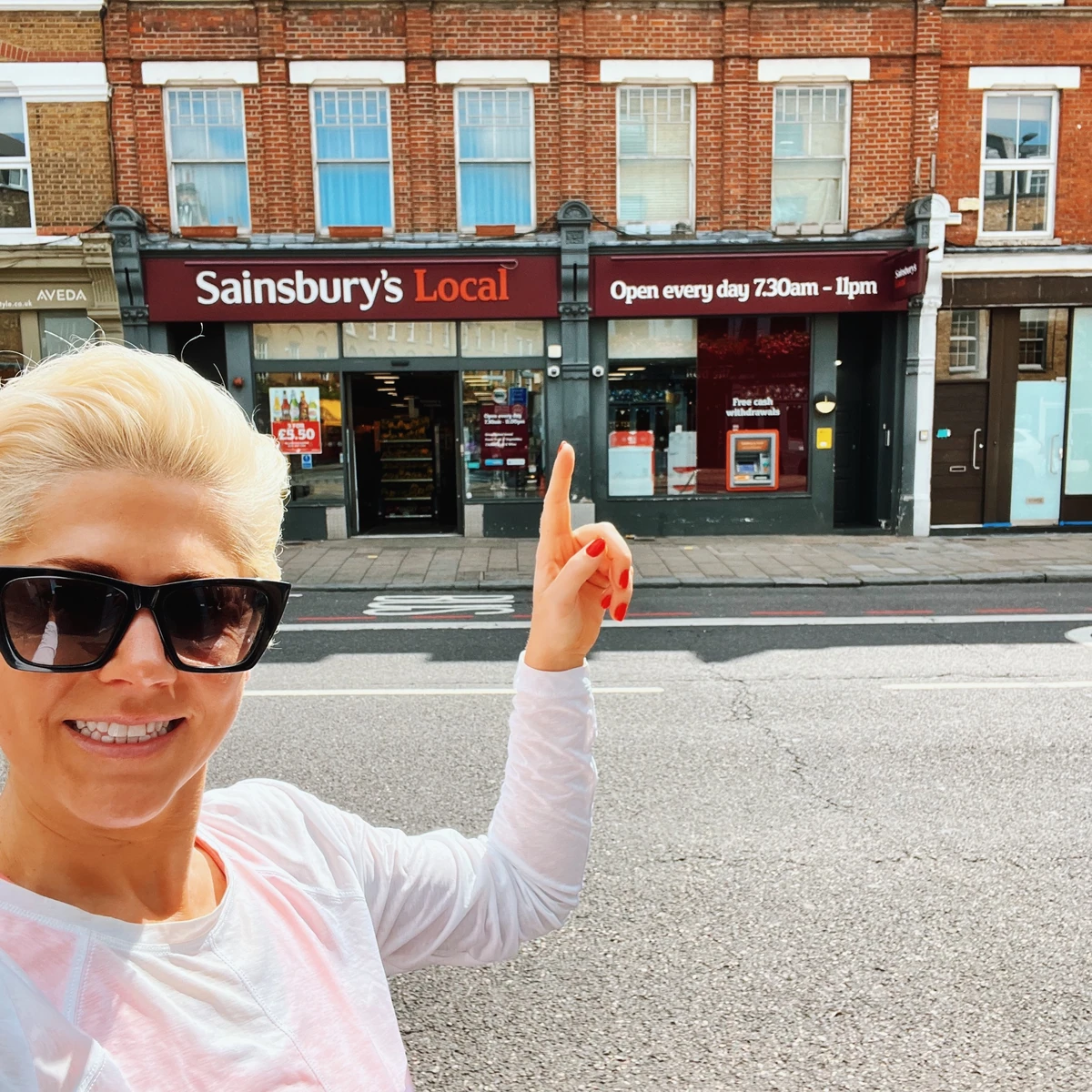 Woman pointing towards a Sainsbury's Local shopfront displaying opening hours