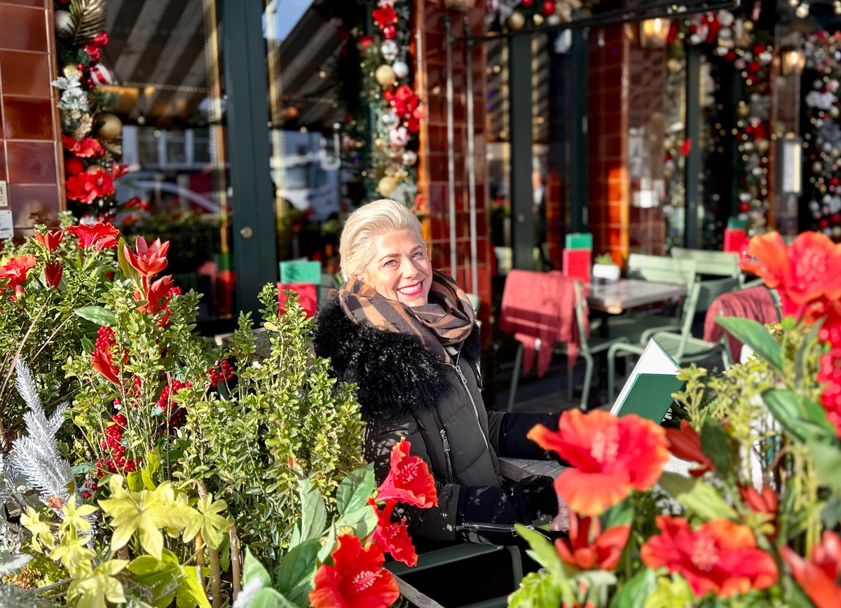 Smiling woman seated outside a festive restaurant, surrounded by vibrant red flowers