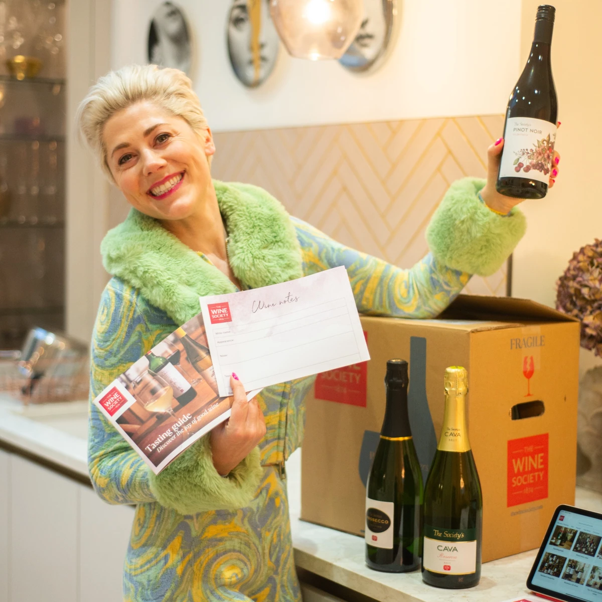 Smiling woman holding a Pinot Noir bottle and wine tasting materials beside wine boxes and bottles