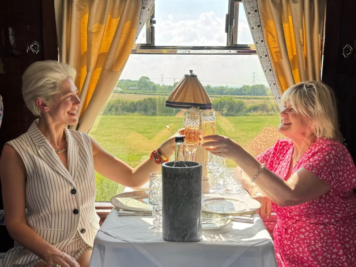 Two women clinking wine glasses aboard a train overlooking countryside fields
