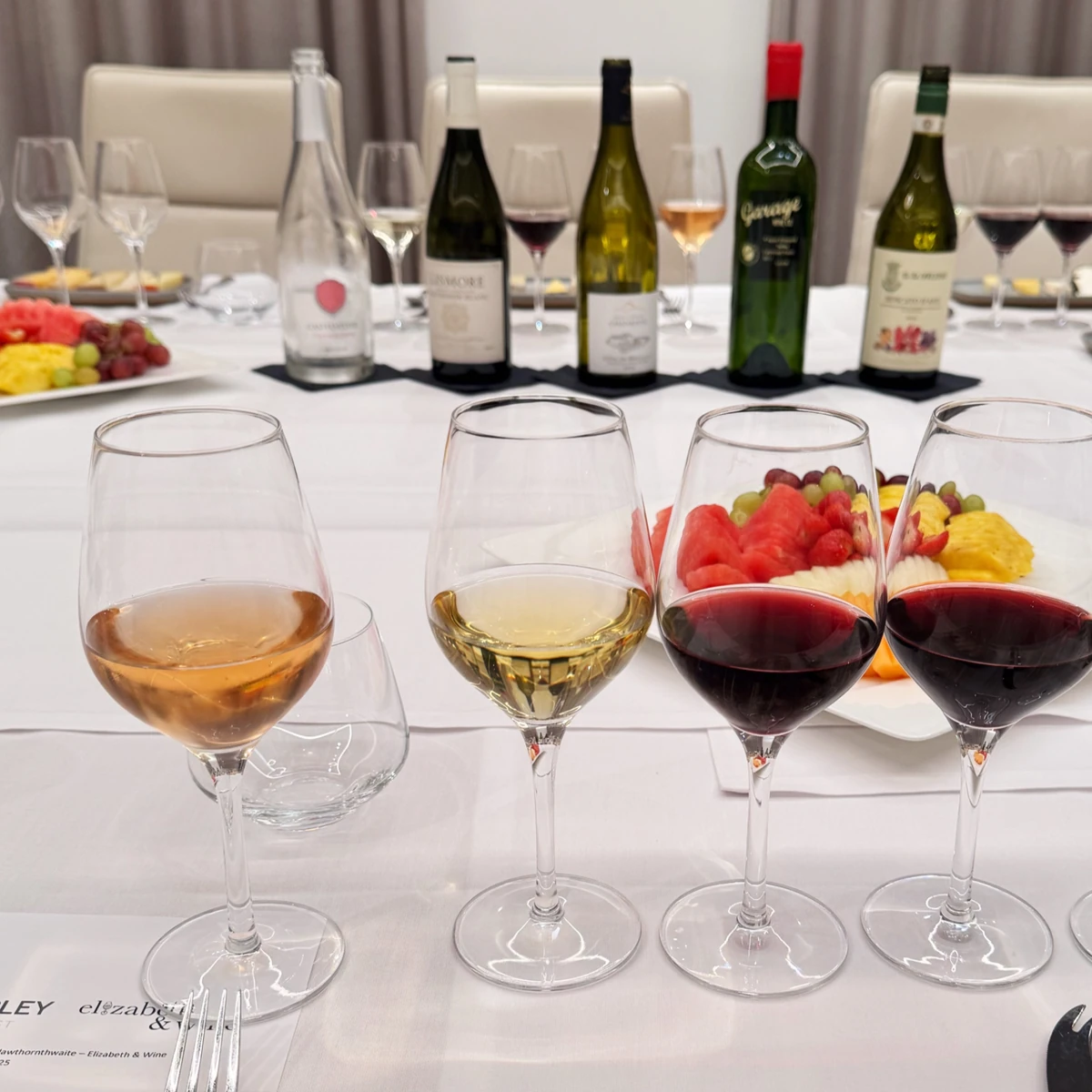 Four wine glasses and five bottles on a dining table with fruit platters