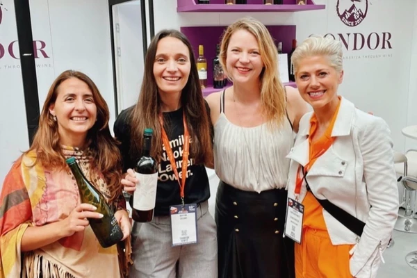 Four women smiling at a wine stand, holding bottles and wearing event badges