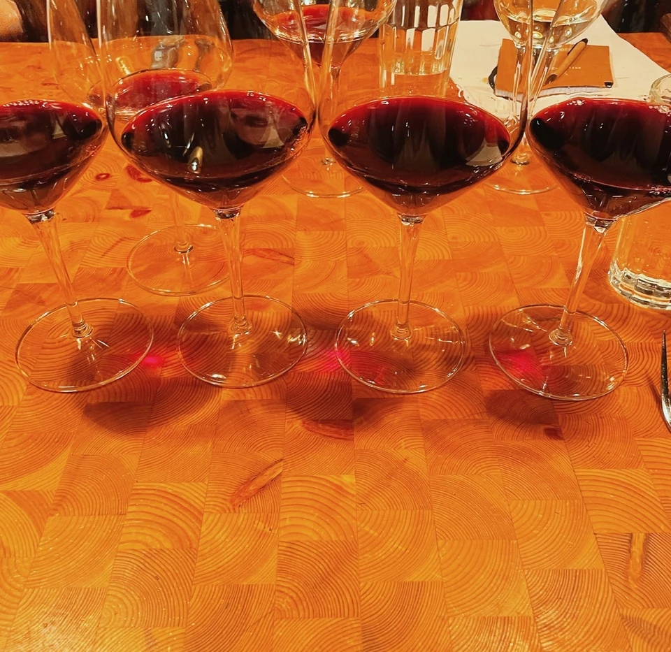 Four glasses of red wine on a wooden tasting table with empty glasses and a water tumbler behind