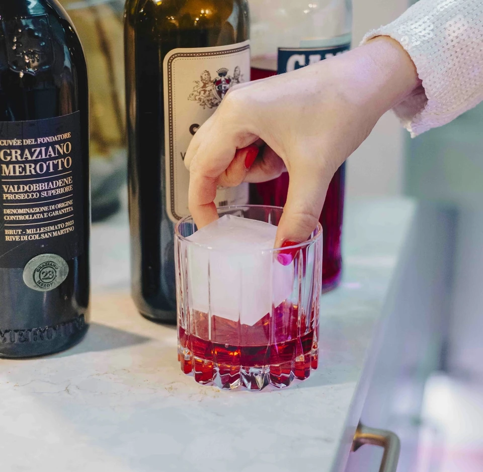 Hand adding a red garnish to a pink Prosecco cocktail beside bottles labelled Valdobbiadene