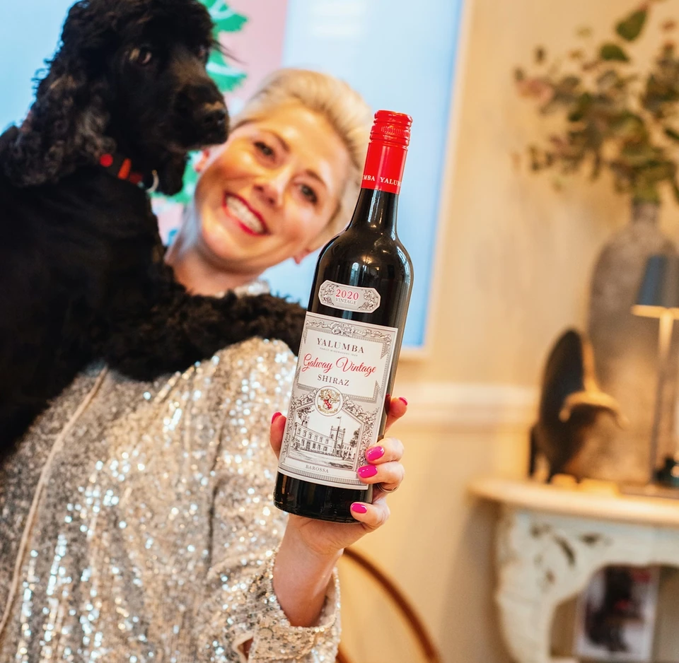 Woman in a sequinned top holds a bottle of Shiraz beside a black dog