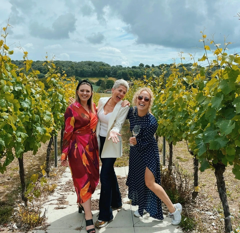 Three women smiling between rows of Hambledon grapevines, with one holding a wine glass