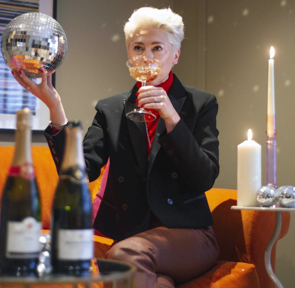 Woman in a black jacket holding a disco ball and toasting with sparkling wine beside candles