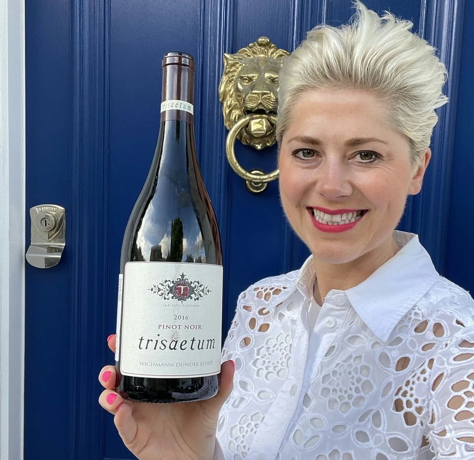 Smiling woman holding a Trisaetum Pinot Noir bottle outside a blue door
