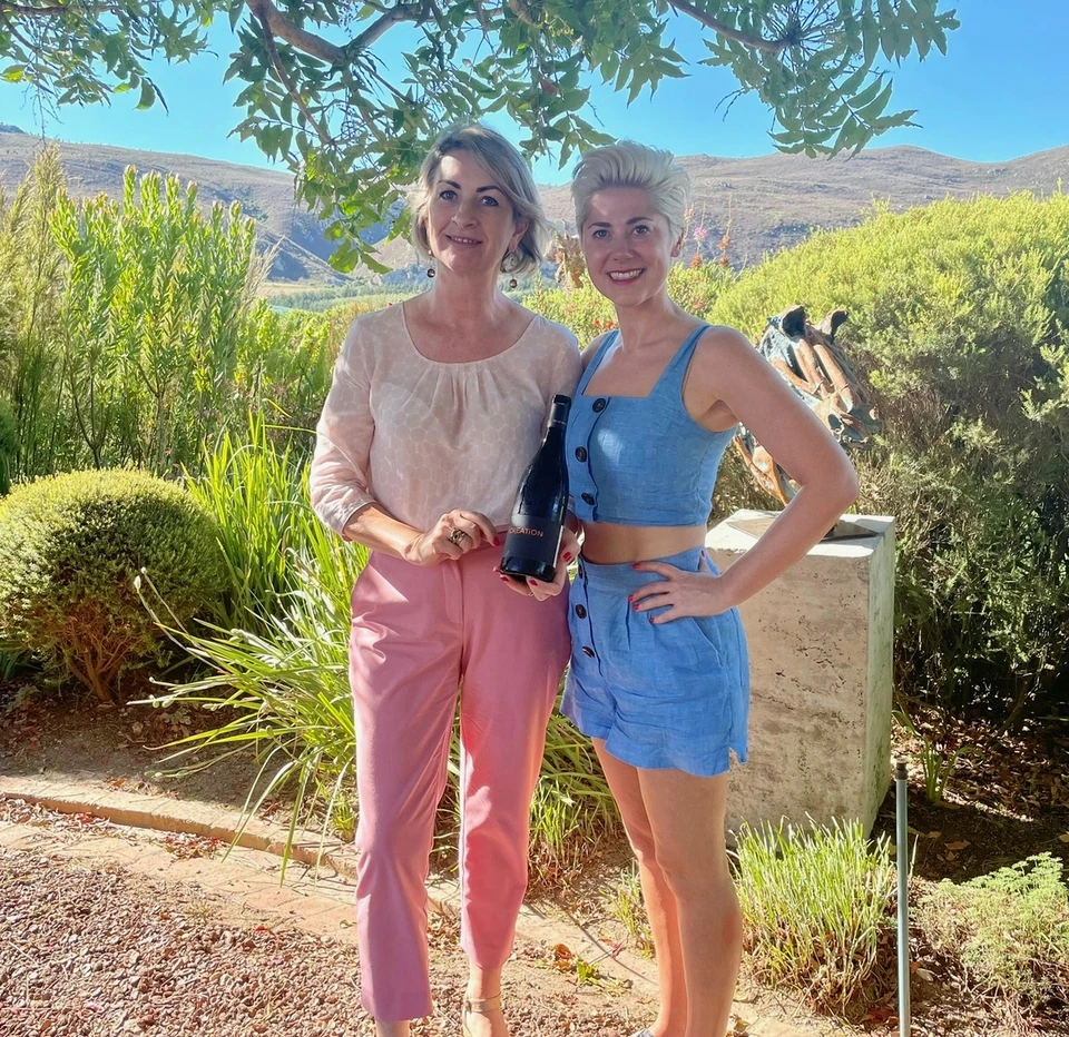Two women pose beneath a tree, one holding a wine bottle, with hills and greenery behind them