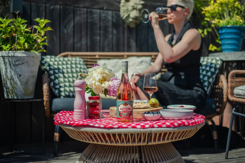 Woman drinking rosé beside a table set with Les Dauphins wine, cheese and fruit