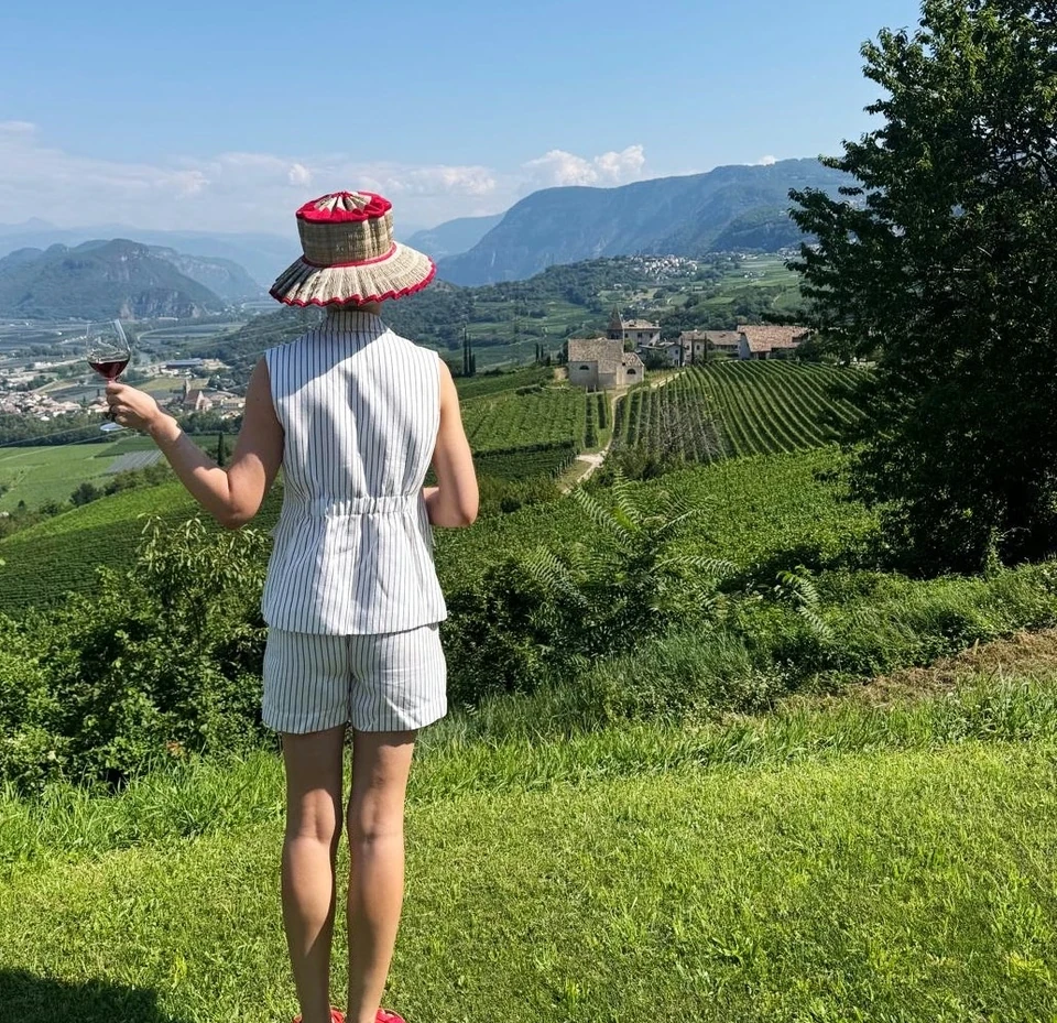 Person in a striped outfit and sun hat raises a wine glass above Alto Adige vineyards and mountains