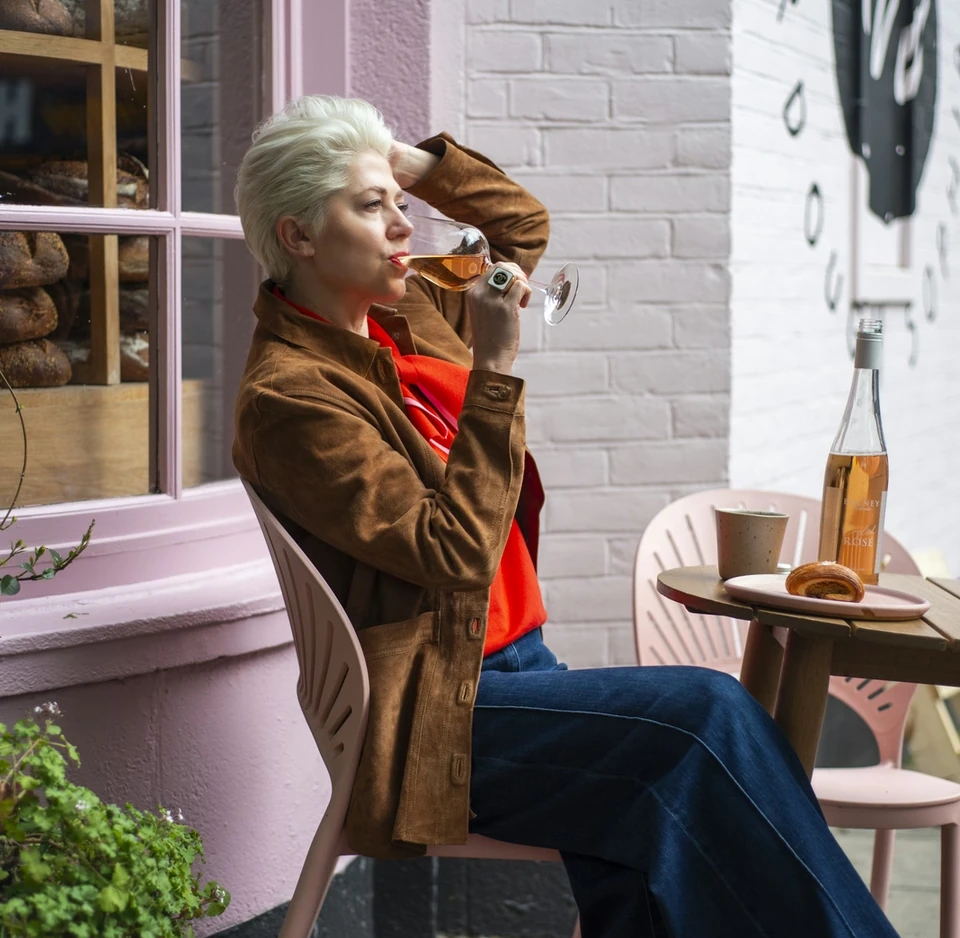 Woman drinking rosé outside a pink-painted café window, with a bottle and pastries on the table