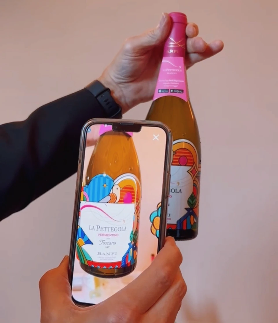 A hand holds a La Pettegola Vermentino bottle while another person photographs the label