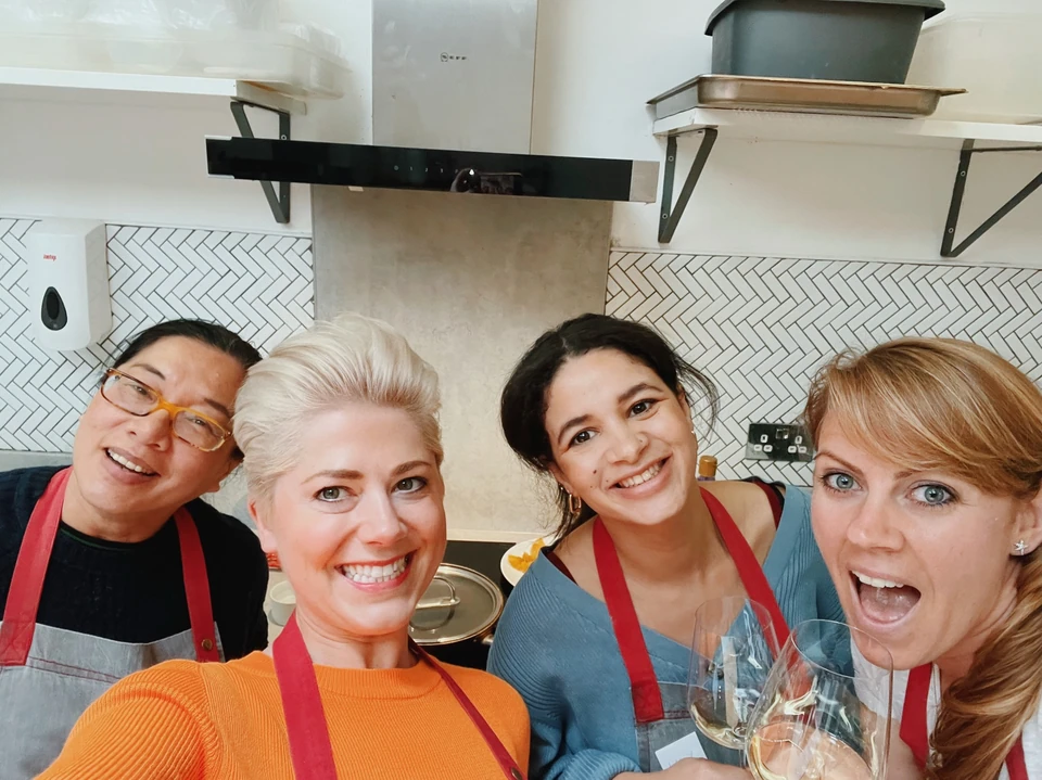 Four women in red aprons pose in a kitchen with glasses of white wine