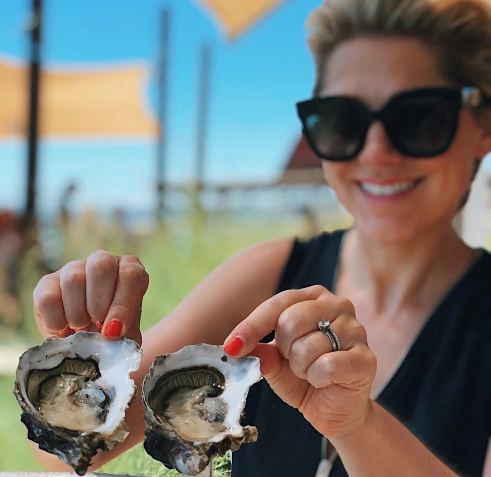 Woman in sunglasses holds two freshly opened oysters at an outdoor Picpoul tasting