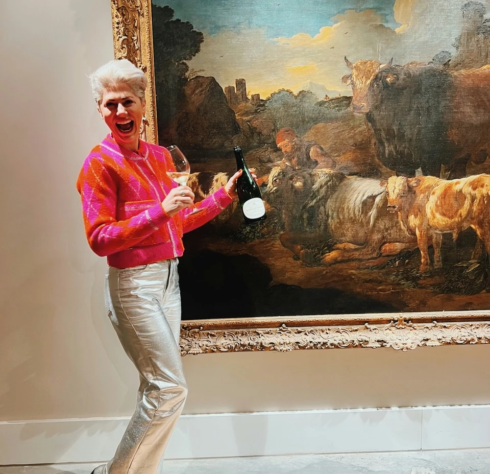 Person holding a wine glass and bottle beside a large framed painting of cattle