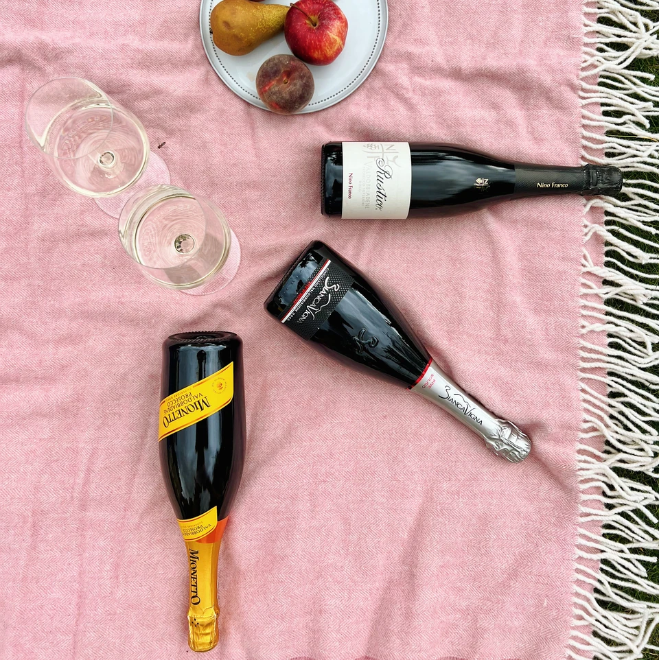 Three Prosecco bottles, two tasting glasses and fruit arranged on a pink blanket outdoors