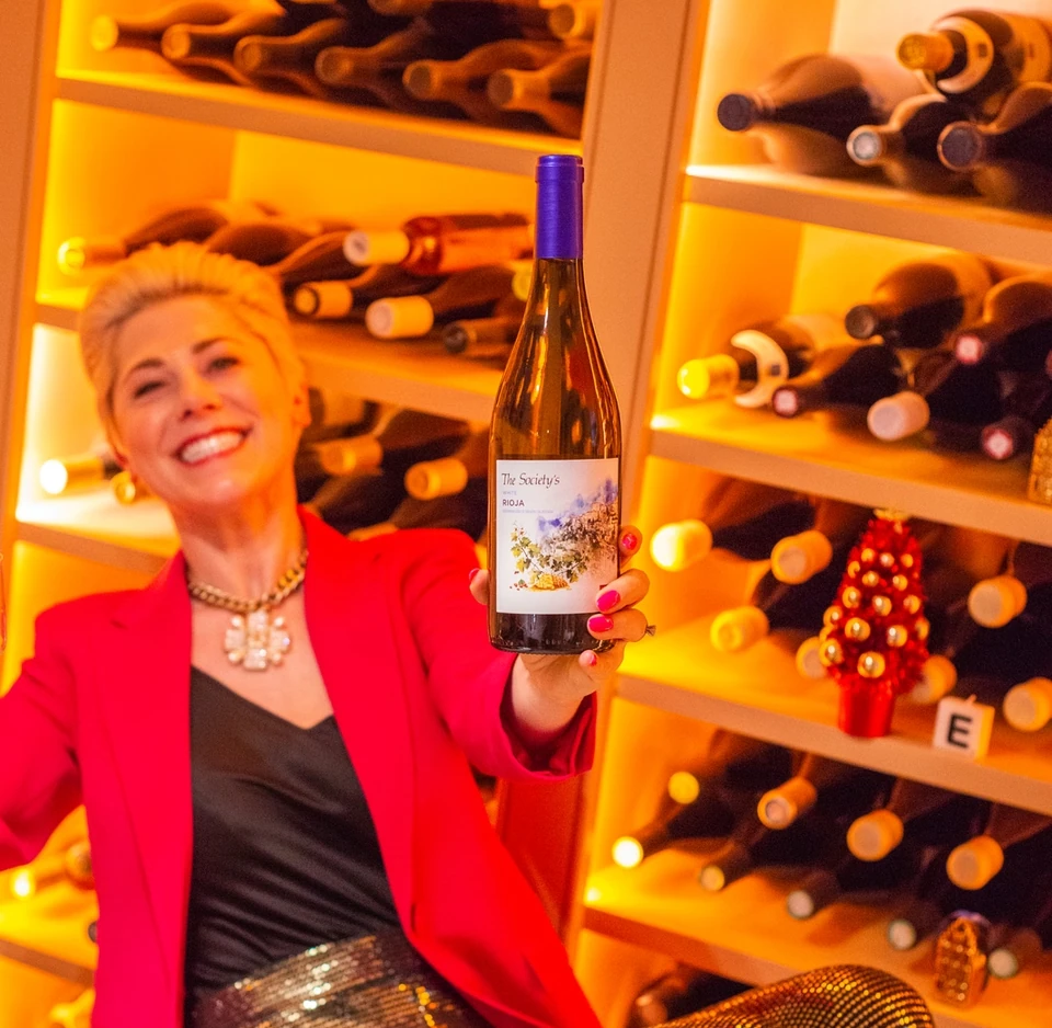 Woman in a red jacket holds a bottle of Rioja in a warmly lit wine cellar