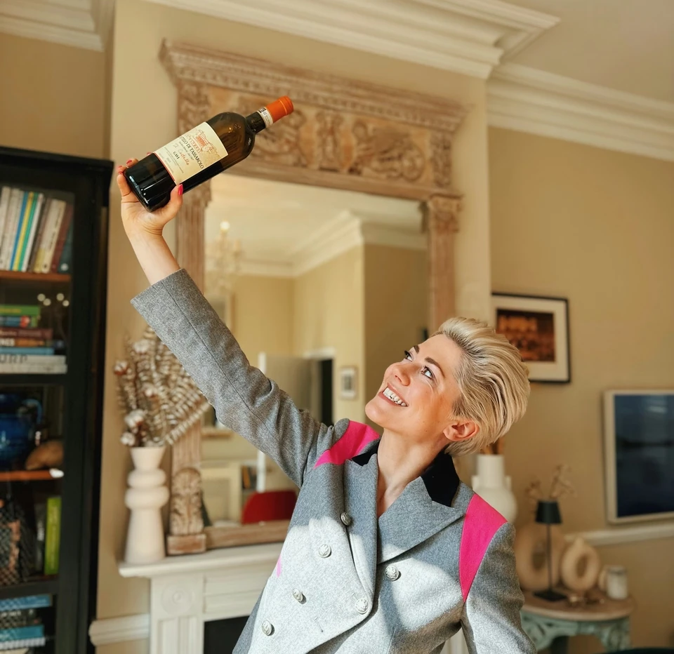 Woman in a grey suit holds a bottle of Gavi wine aloft in an elegant sitting room