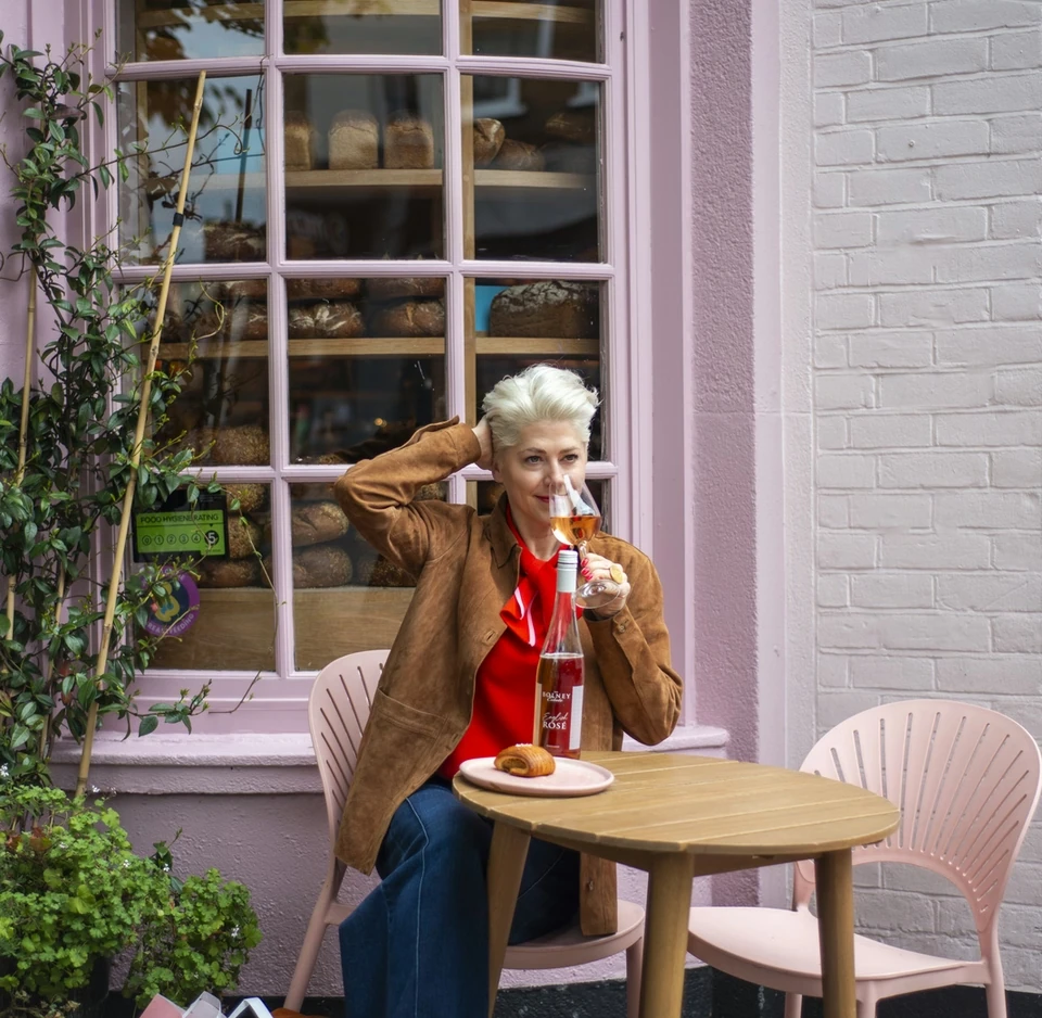 Woman drinking rosé outside a bakery window with a bottle and pastry on the table.