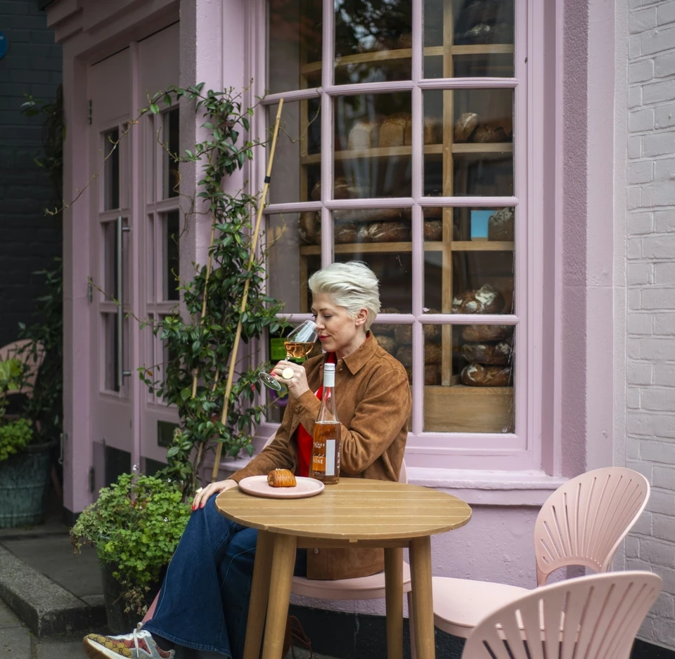 Woman tasting Bolney rosé beside a pink café window, with a pastry and bottle on the table