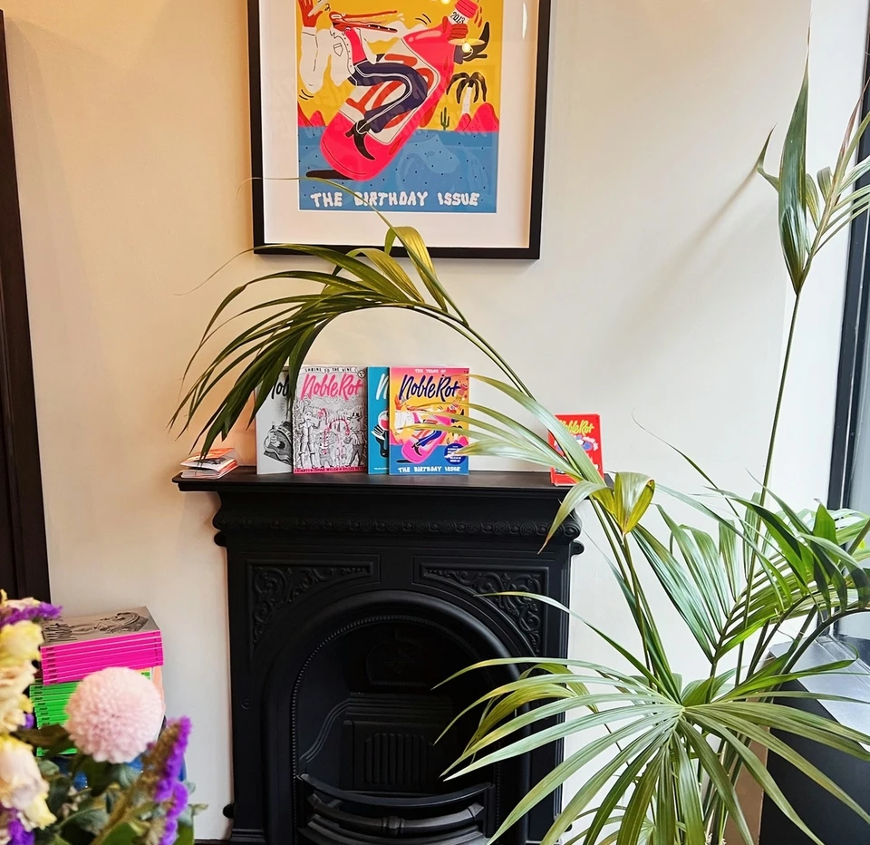 Noble Rot birthday issue framed above magazines, a black fireplace and leafy houseplants