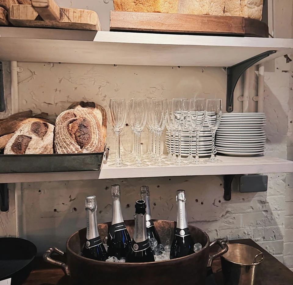 Sparkling wine bottles beside bread, champagne flutes and stacked plates on wooden shelves