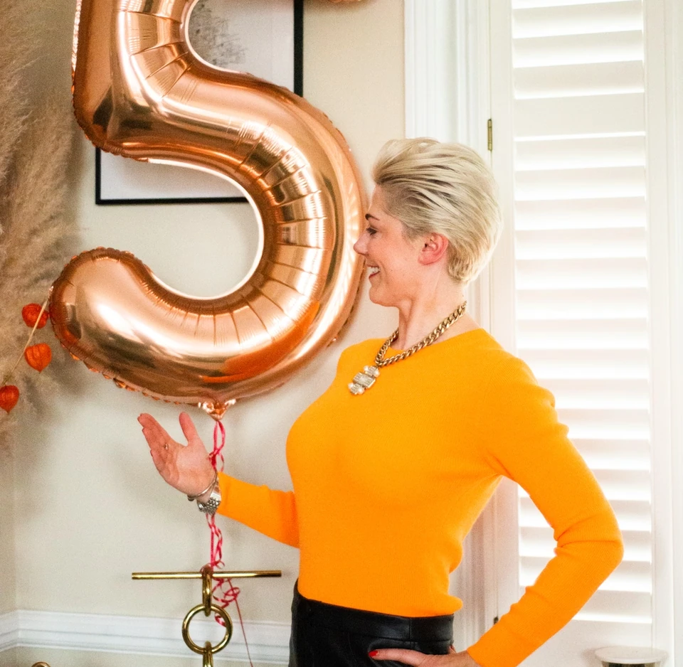 Woman in a bright orange jumper beside a large rose-gold number five balloon