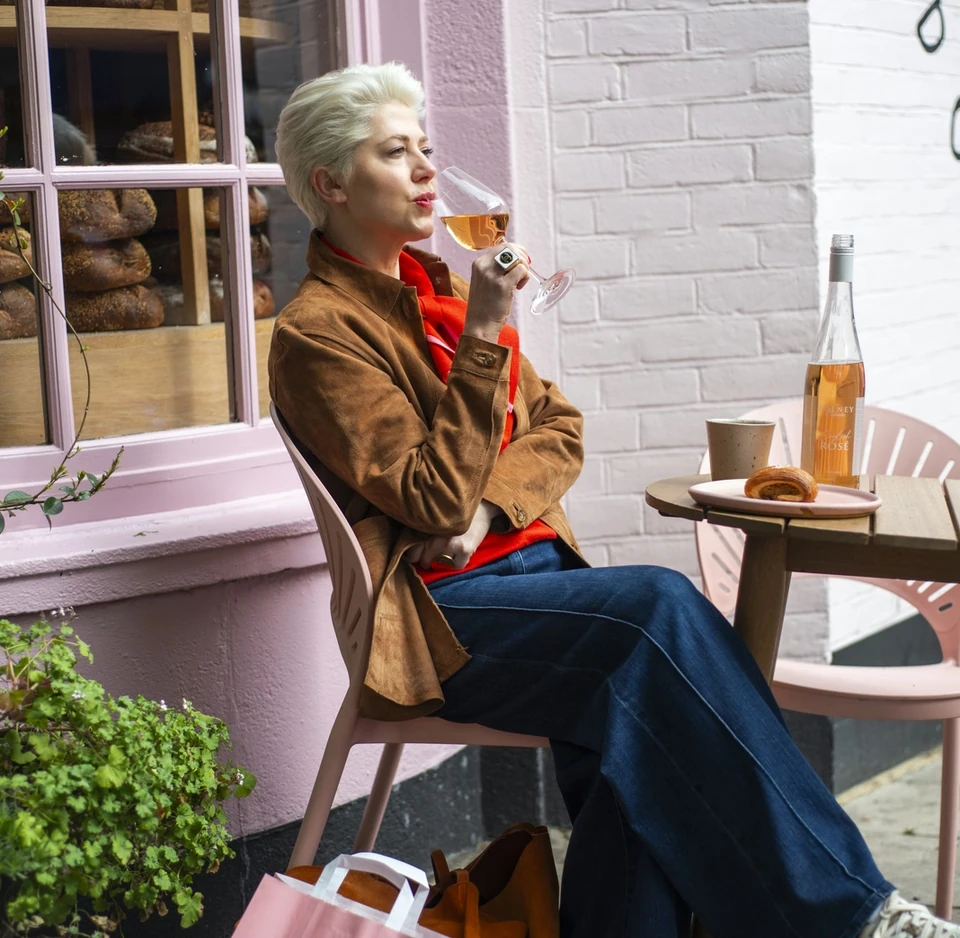 Person tasting rosé beside a bakery window, with a bottle, pastry and coffee on a table